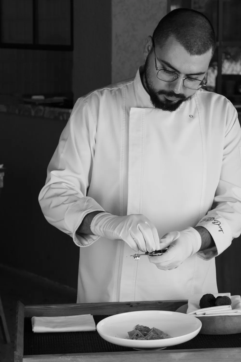 Serhat Doğramacı beyaz aşçı ceketiyle bir tabağın üzerine eğilmiş, elinde rende; siyah-beyaz fotoğraf